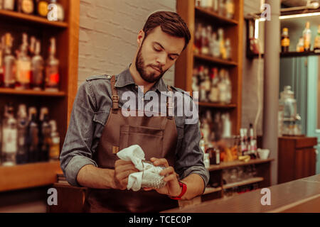 Occhiali di lucidatura. Bello focalizzato duro lavoro di calma dai capelli scuri barbuto giovani-adulti nice-working barman indossando maglietta di cotone e grembiule nero poli Foto Stock