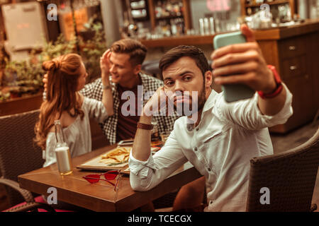 Scattare le foto. Sconvolto pessimista triste annoiato a pelo corto bella barbuto maschio nel suo 25s tenendo selfies mentre i suoi compagni cuddling in background Foto Stock