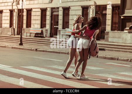 Camminando per le strade con gli amici. Abbagliante bionda dai lunghi capelli radiante sorridente gradevoli bella signora giovane abbracciando con la sua buona-Afro-Am accattivante Foto Stock