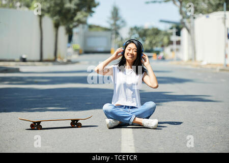 Abbastanza felice Asian ragazza seduta sulla strada accanto a skateboard e ascolto di musica in lei le cuffie Foto Stock