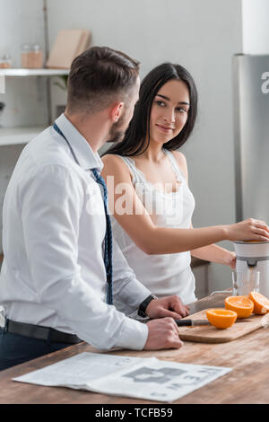 Attraente brunette donna che guarda l'uomo in tuta mentre la spremitura di arance in centrifuga Foto Stock