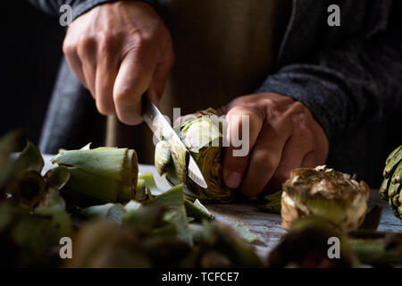 Primo piano di un uomo caucasico di taglio con un coltello da cucina alcune materie carciofi freschi, su un bianco tavola in legno rustico Foto Stock