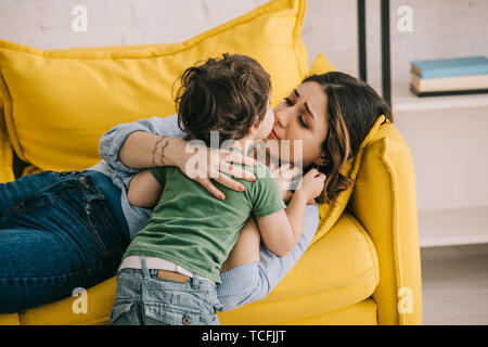 Little Boy kissing stanco madre sdraiato sul divano giallo Foto Stock