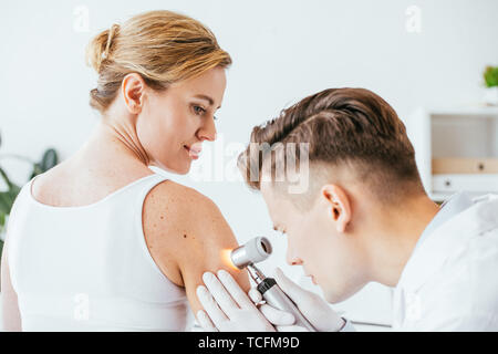 Il dermatologo in guanti in lattice azienda dermatoscope mentre esaminando bella donna con la malattia della pelle Foto Stock