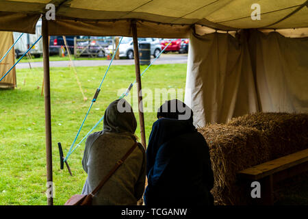 Giovane, sia indossando felpa con cappuccio umido tops, seduto alla porta di una tenda durante un evento, si ripara dalla pioggia. Oltre il punto di vista di spalla. Foto Stock