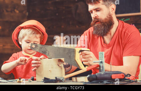 Il ragazzo, bambino impegnato nel casco protettivo di imparare a usare handsaw con papà. Dazi maschile concetto. Padre, genitore con barba insegnamento piccolo figlio di segare con sharp handsaw, falegname artigianato. Foto Stock