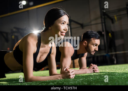 Messa a fuoco selettiva di allegra donna sorridente mentre facendo il listone esercizio vicino all uomo sull'erba Foto Stock