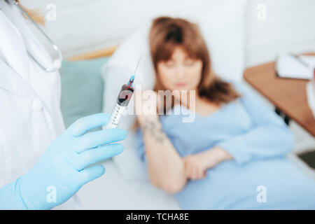 Vista ritagliata del medico tenendo la siringa con il campione di sangue del donatore e giacente sul letto Foto Stock