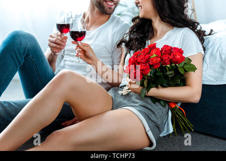 Vista ritagliata della coppia sorridente tintinnanti con bicchieri di vino rosso e la donna azienda rose rosse Foto Stock