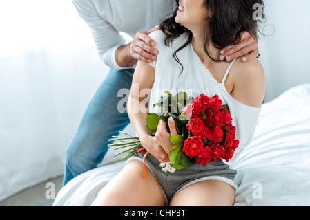 Vista ritagliata dell'uomo abbracciando donna sorridente con bouquet di rose rosse Foto Stock