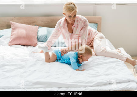 Allegro madre bionda guardando simpatico bimbo figlio di strisciare sul letto Foto Stock