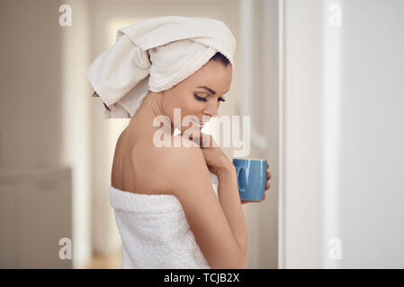 Bella donna di mezza età avvolto nelle fresche e pulite asciugamani bianchi attorno alla sua testa e corpo appoggiato contro una parete interna con una tazza di bevanda in un cost Foto Stock