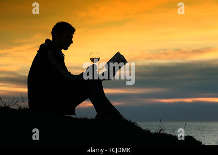 Silhouette Ragazzo seduto sulla scogliera in sera vicino al mare, legge libro e bere un bicchiere di vino Foto Stock