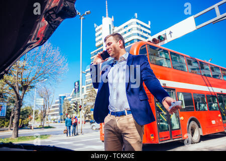 Giovane imprenditore sta parlando sul suo telefono cellulare mentre guardando sotto il cofano sollevato della sua vettura. Foto Stock