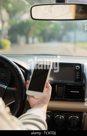 Femmina giovane conducente utilizzando il touch screen smartphone e mano che tiene il volante in un'auto. Foto Stock