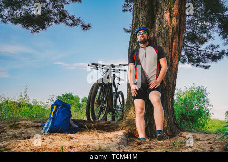 Maschio caucasico ciclista uno stile di vita attivo in appoggio sotto agli alberi dopo esercizio bicicletta acqua potabile dalla bottiglia. Sportivo in casco e abbigliamento sportivo. Ha Foto Stock