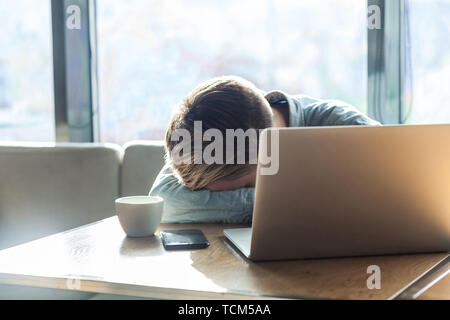 Ritratto di tristezza premuto stanco giovane uomo freelancer in maglietta blu sono seduti da soli in cafe e dormire sul tavolo dopo un duro lavoro e le ore di lavoro straordinario w Foto Stock