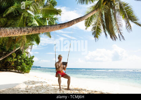 Uomo in vacanza seduta oltre lo Swing guardando a tavoletta digitale presso la spiaggia Foto Stock