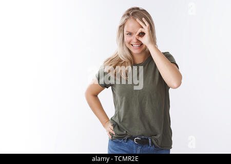 Carismatico felice giovane donna con capelli biondi e occhi blu spiata attraverso il foro che mostra bene gesto su occhio e guardando la telecamera divertito e gioioso gr Foto Stock