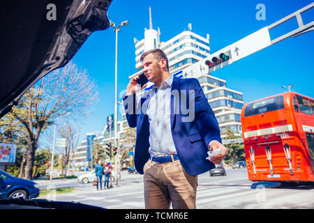 Giovane imprenditore sta parlando sul suo telefono cellulare mentre guardando sotto il cofano sollevato della sua vettura. Foto Stock