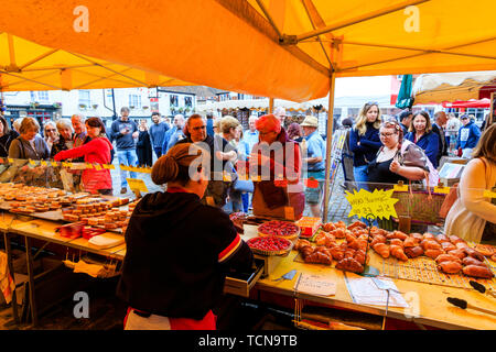 Le Weekend event mercato francese a sandwich, Inghilterra. Donna comprare il pane dal forno Francese stallo, commerciante di mercato che serve. Contatore coperti in vari bastoncini francesi, pane e torte. Foto Stock