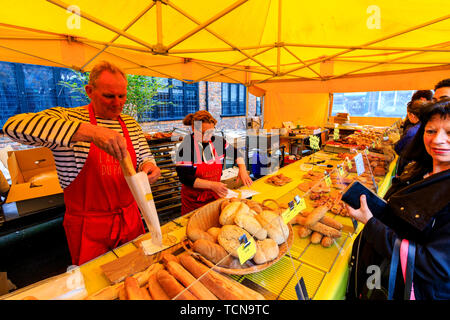 Le Weekend event mercato francese a sandwich, Inghilterra. Donna comprare il pane dal forno Francese stallo, commerciante di mercato che serve, mettendo il pane in borsa. Contatore coperti in vari bastoncini francesi, pane e torte. Foto Stock