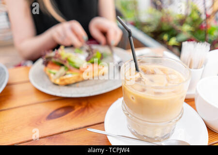 La messa a fuoco in primo piano sul caffè freddo con cubetti di ghiaccio. Una giovane donna è avere la prima colazione in una caffetteria estiva. Foto Stock