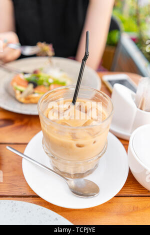 La messa a fuoco in primo piano sul caffè freddo con cubetti di ghiaccio. Una giovane donna è avere la prima colazione in una caffetteria estiva. Foto Stock