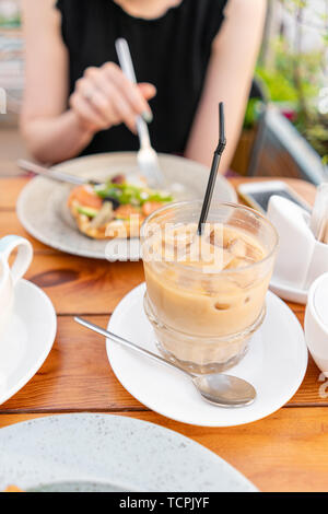 La messa a fuoco in primo piano sul caffè freddo con cubetti di ghiaccio. Una giovane donna è avere la prima colazione in una caffetteria estiva. Foto Stock