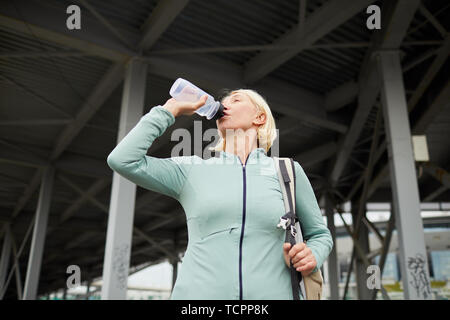 Bionda matura sportive assetato di bere acqua da bottiglie di plastica dopo il duro allenamento in ambiente urbano sul giorno di estate Foto Stock