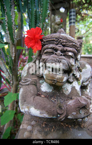Tipico Dvarapala Balinese statua di pietra con un rosso di fiori di ibisco, catturato in un parco in Ubud a Bali, Indonesia Foto Stock
