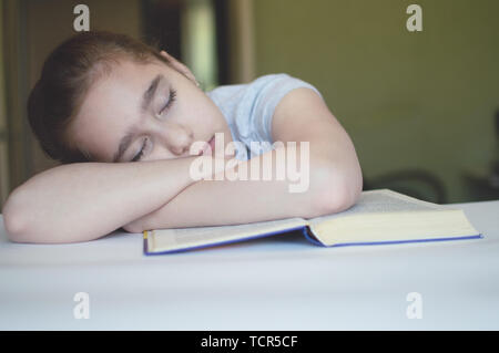 Torturato ragazza bambino legge un libro a tavola e lei è molto stanco e vuole dormire, sbadigli, si addormenta durante la lettura di storie e di lezioni Foto Stock