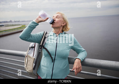 Sete bionda matura donna attiva nell'abbigliamento sportivo di bere acqua da bottiglie di plastica mentre in piedi da riverside Foto Stock