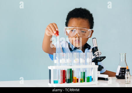 Grave African American Boy in occhiali di protezione utilizzando una pipetta durante il tentativo di imparare a mescolare prodotti chimici colorati in laboratorio Foto Stock