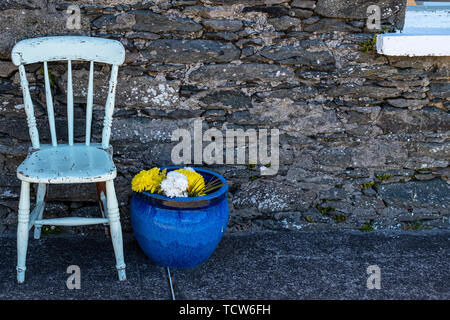 Un bianco verniciato sedia in legno con un grande blu impianto ceramica pentola con giallo e bianco fiori contro un antico in pietra della parete esterna di una casa. Foto Stock