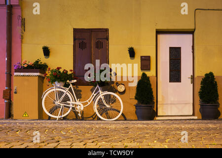 Bianca decorativa bicicletta con cesti di fiori su di esso, un orologio sulla parete nelle vicinanze una bloccato porta bianca e una finestra chiusa. Esterno, scena notturna in Sibiu ( Foto Stock