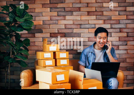 Giovane maschio asiatici imprenditori utilizzare computer portatile e telefono con scatole per offrire ai clienti da ordini - business online e di ecommerce Foto Stock