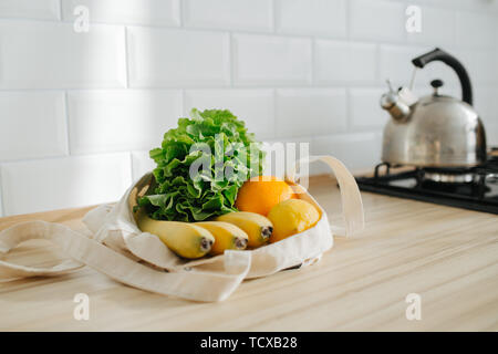 La lattuga, limone, banana e arancio in un bianco borsa di cotone nella soleggiata cucina, giacente sul tavolo. Foto Stock