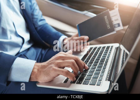 Il check-in. Immagine ritagliata del giovane imprenditore nel formale di lavoro di usura sul laptop e azienda passaporto con biglietti di volo stando seduti in auto. Travel co Foto Stock