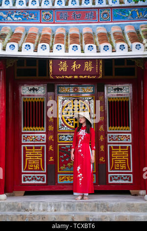 Una giovane donna vietnamita che indossa un abito tradizionale Ao dai e si erge fuori da un tempio nel centro storico della città, Hoi An, Vietnam Foto Stock