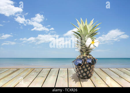 Ananas con occhiali da sole e fiore su pavimento di legno accanto alla spiaggia. Foto Stock
