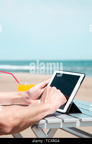 Primo piano di un giovane uomo caucasico sulla spiaggia, utilizzando una tavoletta digitale, seduti a un tavolo all'aperto, accanto a un bicchiere con una fresca bevanda a base di arancia, un Foto Stock