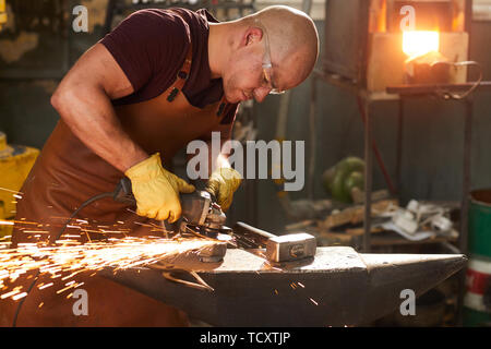 Concentrato occupato giovane operaio in occhiali di sicurezza e grembiule in pelle di metallo di lucidatura con la fresa a incudine in officina Foto Stock