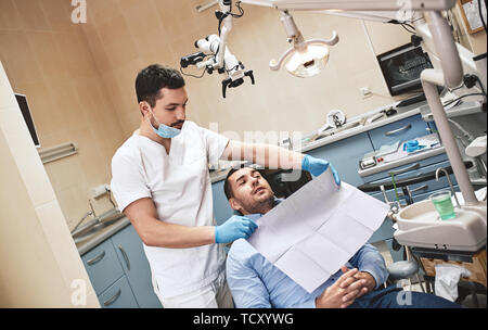 Barbuto dai capelli scuri igienista dentale indossando bianco uniforme è pronto per controllare i denti del paziente. Egli si erge nei pressi di poltrona odontoiatrica a parlare con il suo paziente Foto Stock