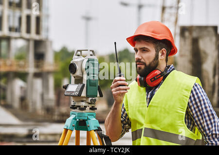 Misurazioni precise. Ingegnere geometra in abbigliamento protettivo e casco rosso utilizzando apparecchiature geodetici e parlando da walkie talkie durante il riposo a cons Foto Stock