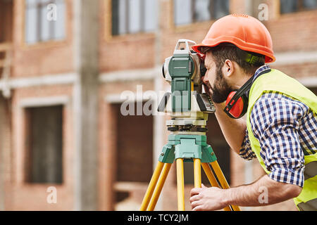 Rilevamenti geodetici. Ingegnere geometra in abbigliamento protettivo e casco rosso utilizzando apparecchiature geodetiche al sito in costruzione. Attrezzature professionali. Costruzione di co Foto Stock