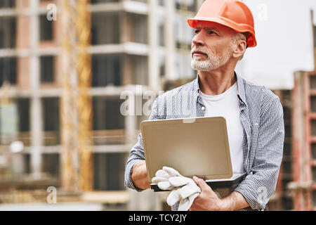 Lavorare all'esterno. Ritratto di occupato costruzione senior engineer in abbigliamento protettivo utilizzando laptop e guardando al processo di lavoro durante la visita a palazzo Foto Stock