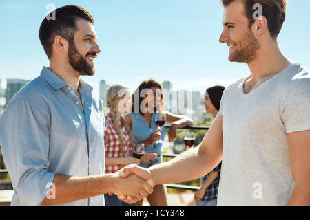 Piacere di conoscerti! Due giovani felici uomini stringono le mani e sorridenti mentre si sta in piedi sul tetto con gli amici. Concetto di barbecue. Concetto di estate Foto Stock