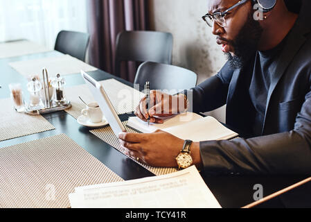 Elegante gravi giovani Afro American businessman in blu scuro giacca e occhiali seduti a tavola, interni che indossa gli auricolari wireless, chiacchierando con Foto Stock