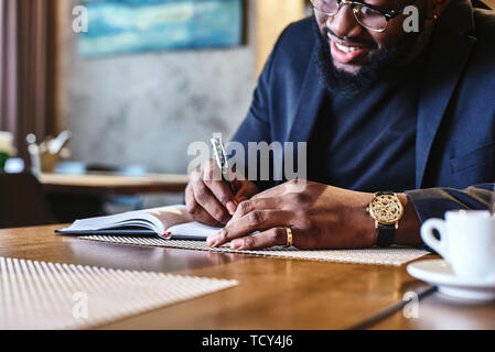 Close up di eleganti giovani afro-americano di CEO in blu scuro giacca passando attraverso alcuni documenti, seduti al tavolo del bar con i documenti e la tazza di caffè Foto Stock